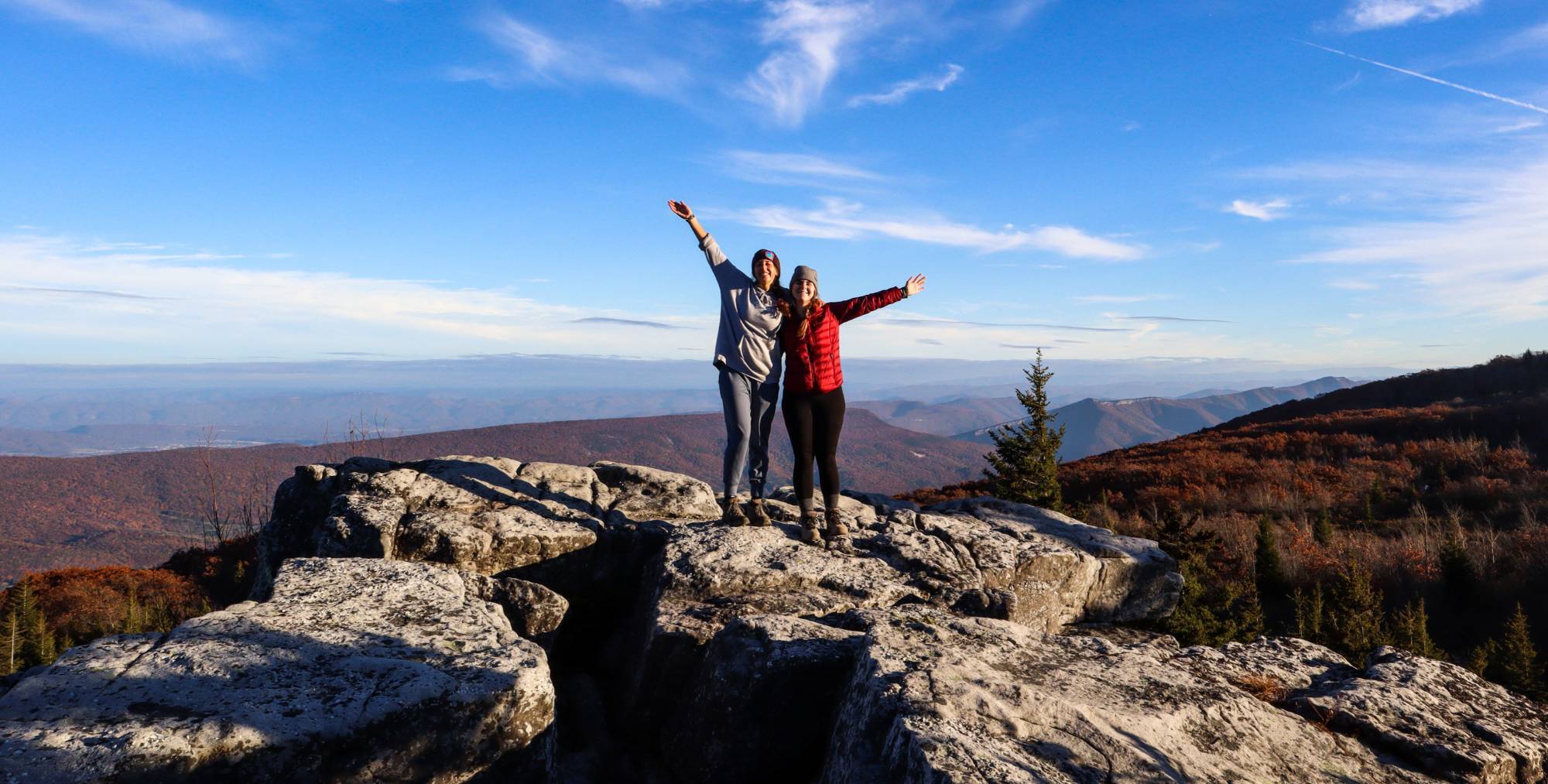 two students standing at the edge of a rocky overlook with arms raised in the air with a mountain range in the distance